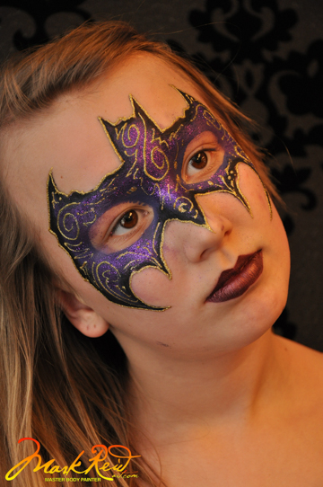 girl with a mask painted around her eyes in the shape of a bat in purple and gold